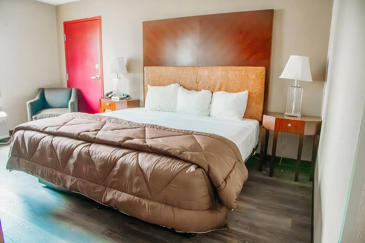A hotel room with a large bed, a wooden headboard, a red door, a chair, a nightstand with a lamp, and a bedside table with another lamp.