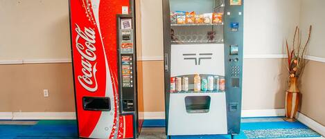 Dos máquinas expendedoras, una de Coca-Cola y otra con un logotipo genérico, en una habitación con alfombra azul y un jarrón con plantas secas.