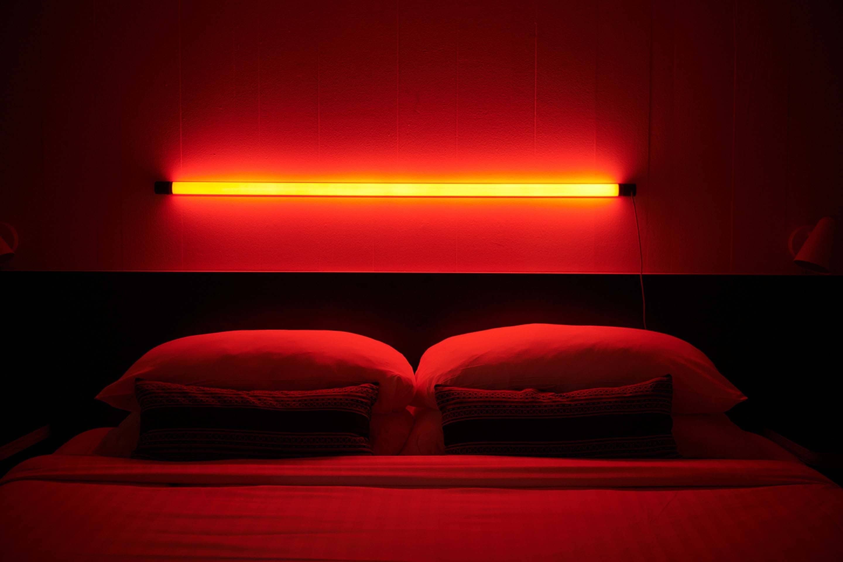 A bed with red bedding and pillows, a red wall, and a horizontal light fixture above the bed.