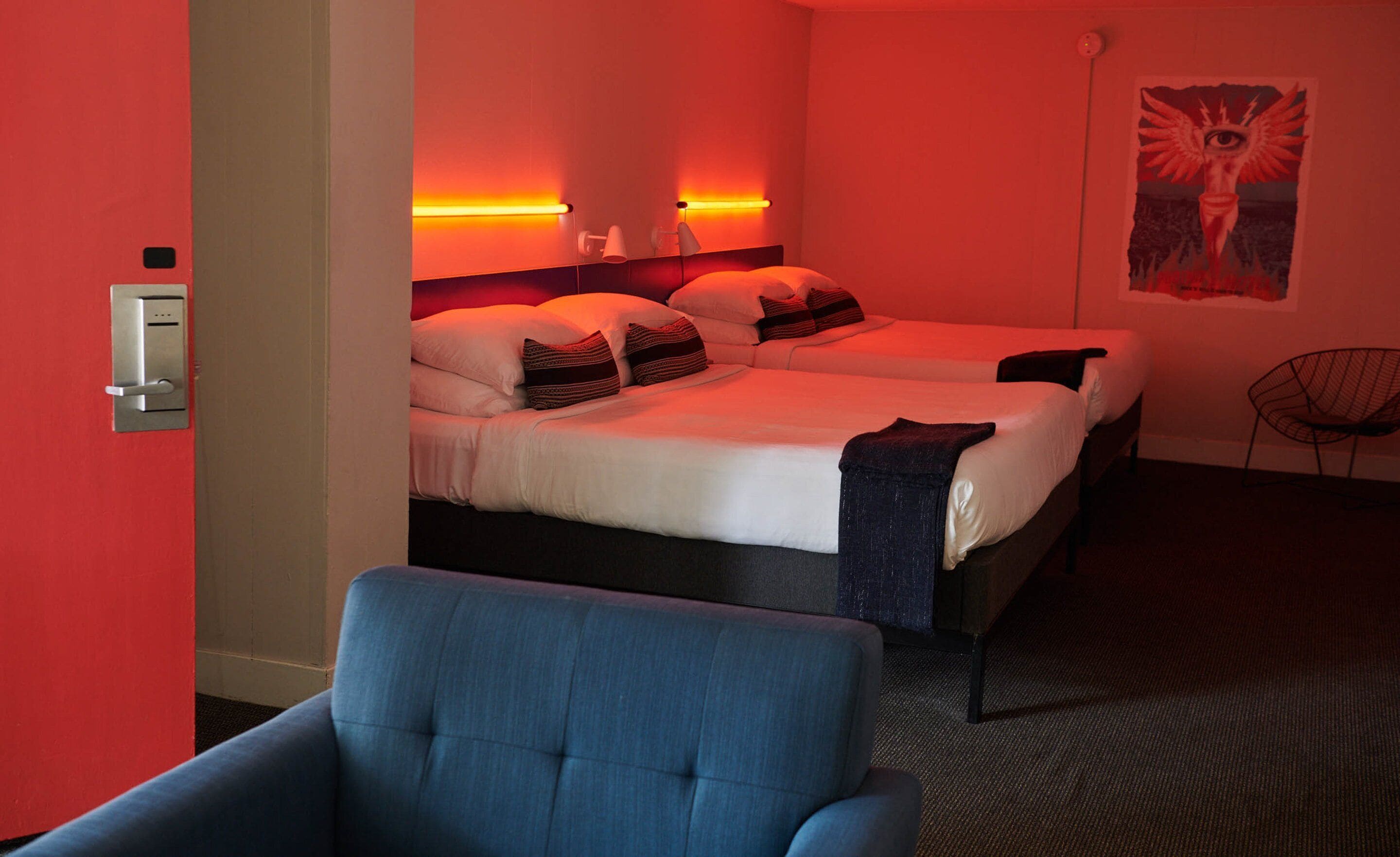 Hotellihuone, jossa on kaksi sänkyä, sininen nojatuoli ja juliste seinällä.