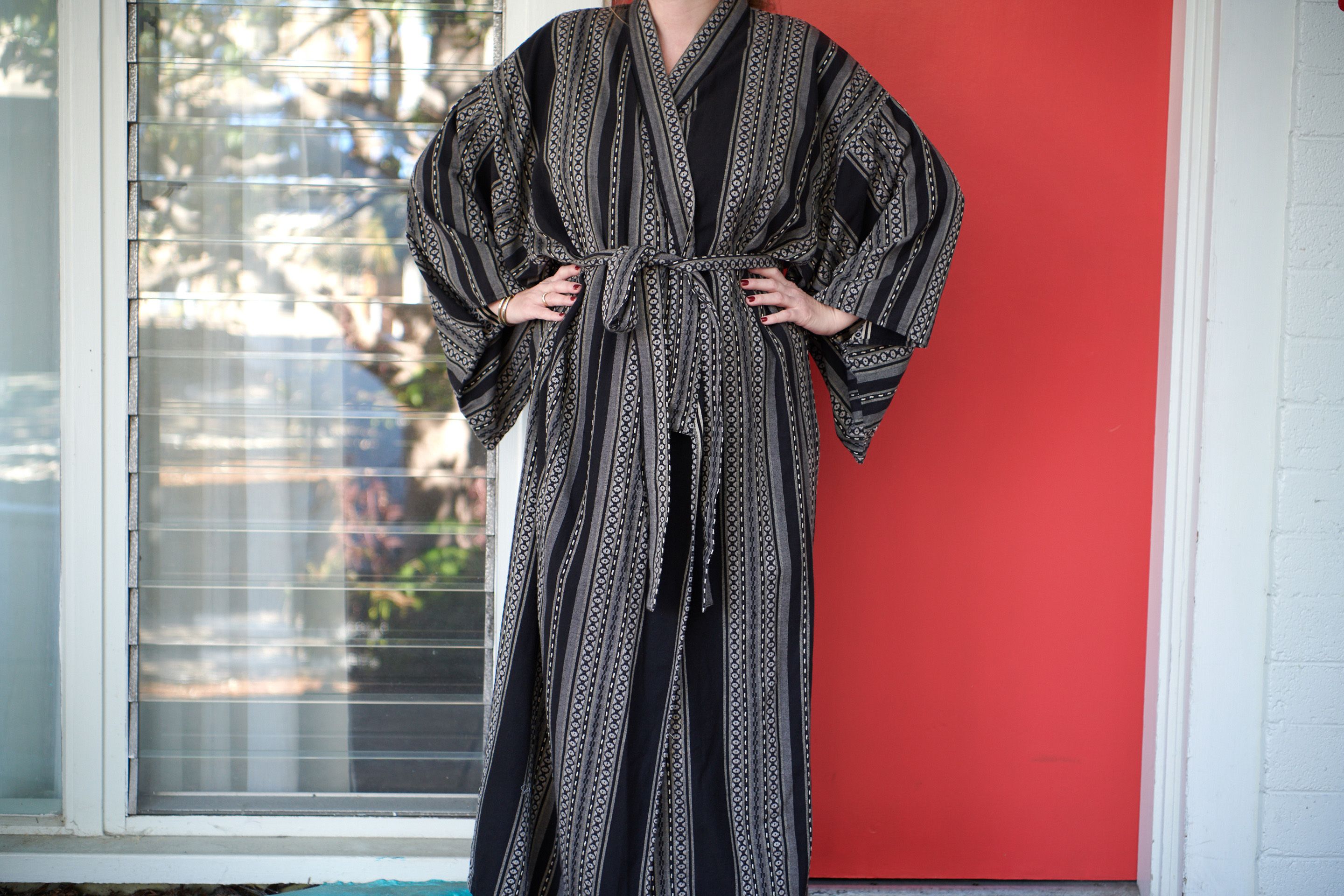 A person standing in front of a window with blinds, wearing a patterned robe.