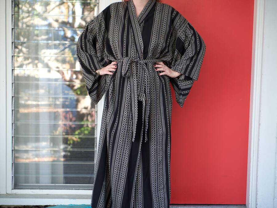 A person standing in front of a window with blinds, wearing a patterned robe.