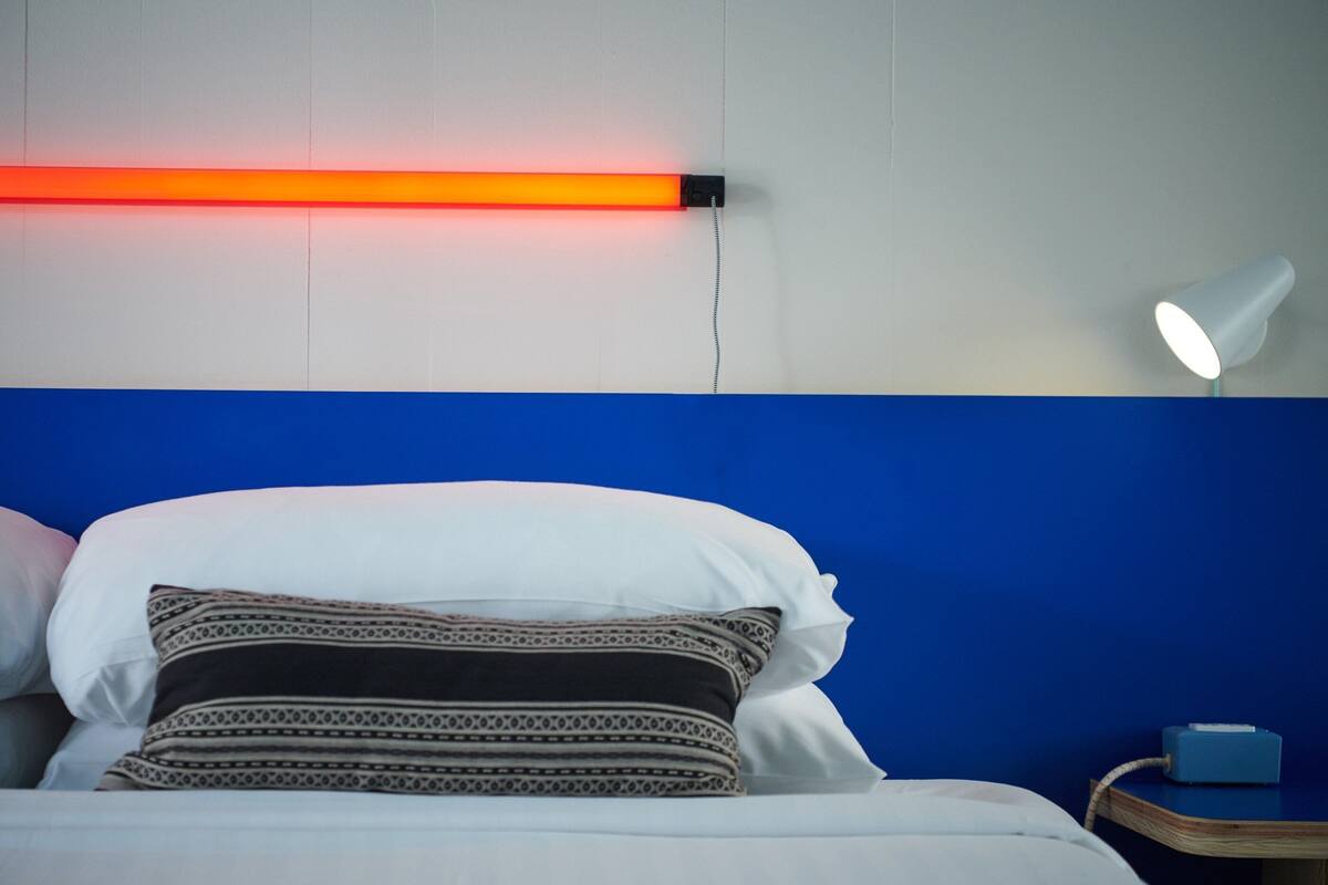 A bed with white and patterned pillows, a blue headboard, a bedside table with a lamp, and a wall-mounted light.