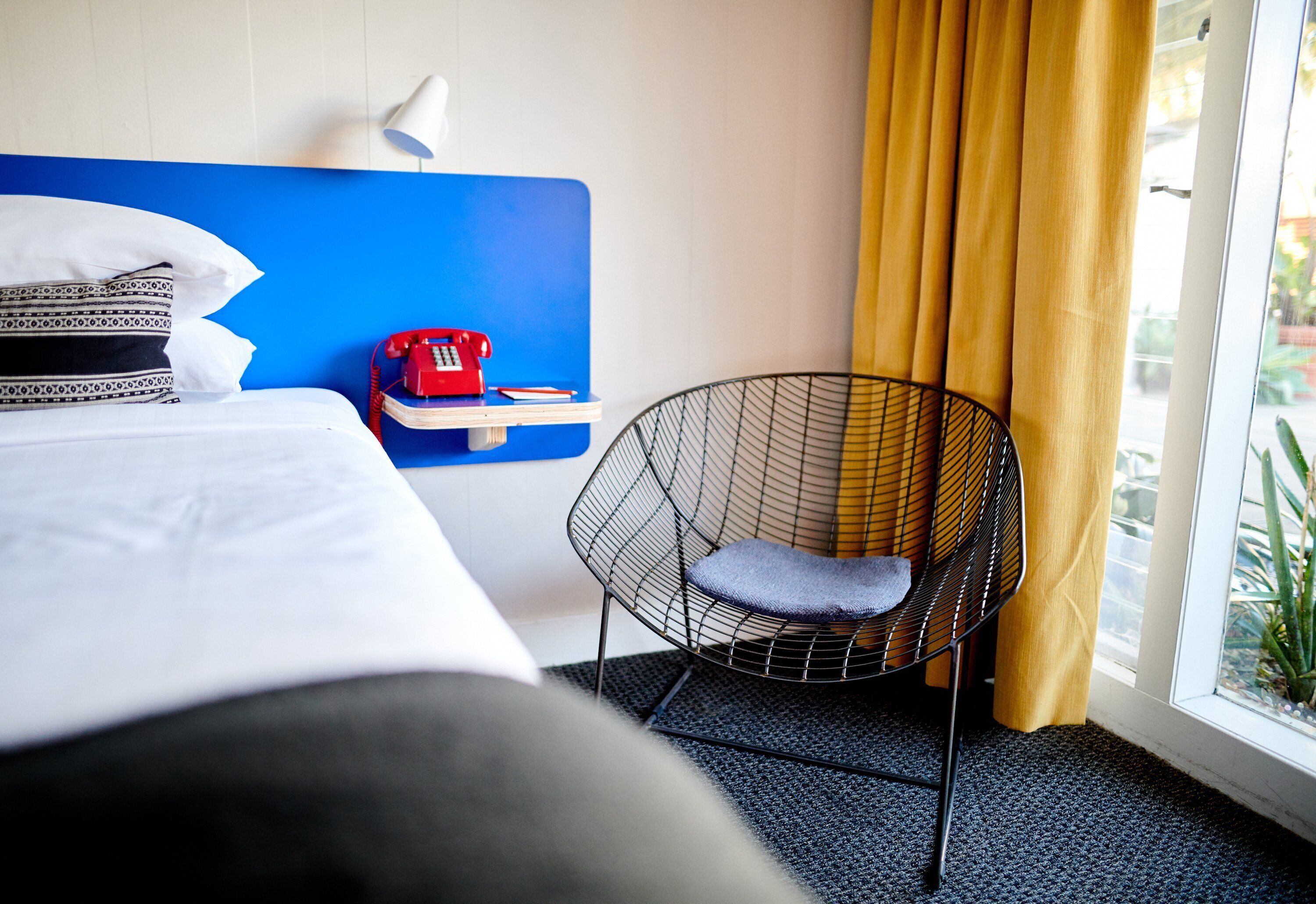 Ein Hotelzimmer mit einem Bett, einem roten Telefon, einem schwarzen Sessel mit Kissen und einem Fenster mit gelben Vorhängen.