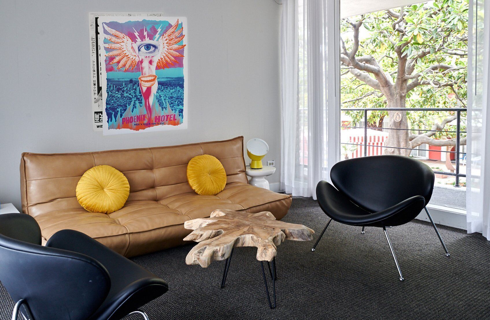 A modern living room with a brown leather sofa, two yellow cushions, a black chair, and a wooden coffee table. There is a colorful poster on the wall and a view of a tree through a window.
