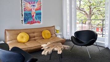 A modern living room with a brown leather sofa, two yellow cushions, a black chair, and a wooden coffee table. There is a colorful poster on the wall and a view of a tree through a window.