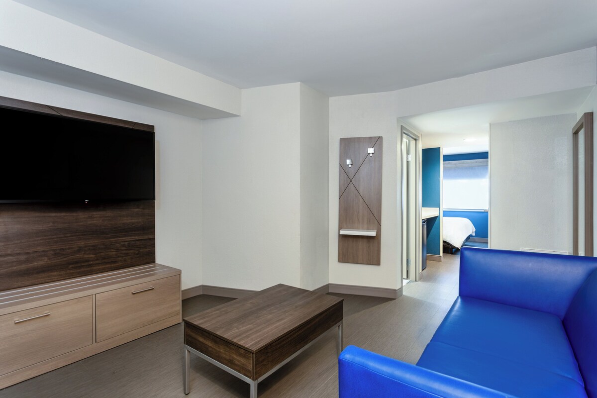 A modern hotel room with a flat-screen TV, a wooden desk, a blue sofa, and a bed with a blue headboard.