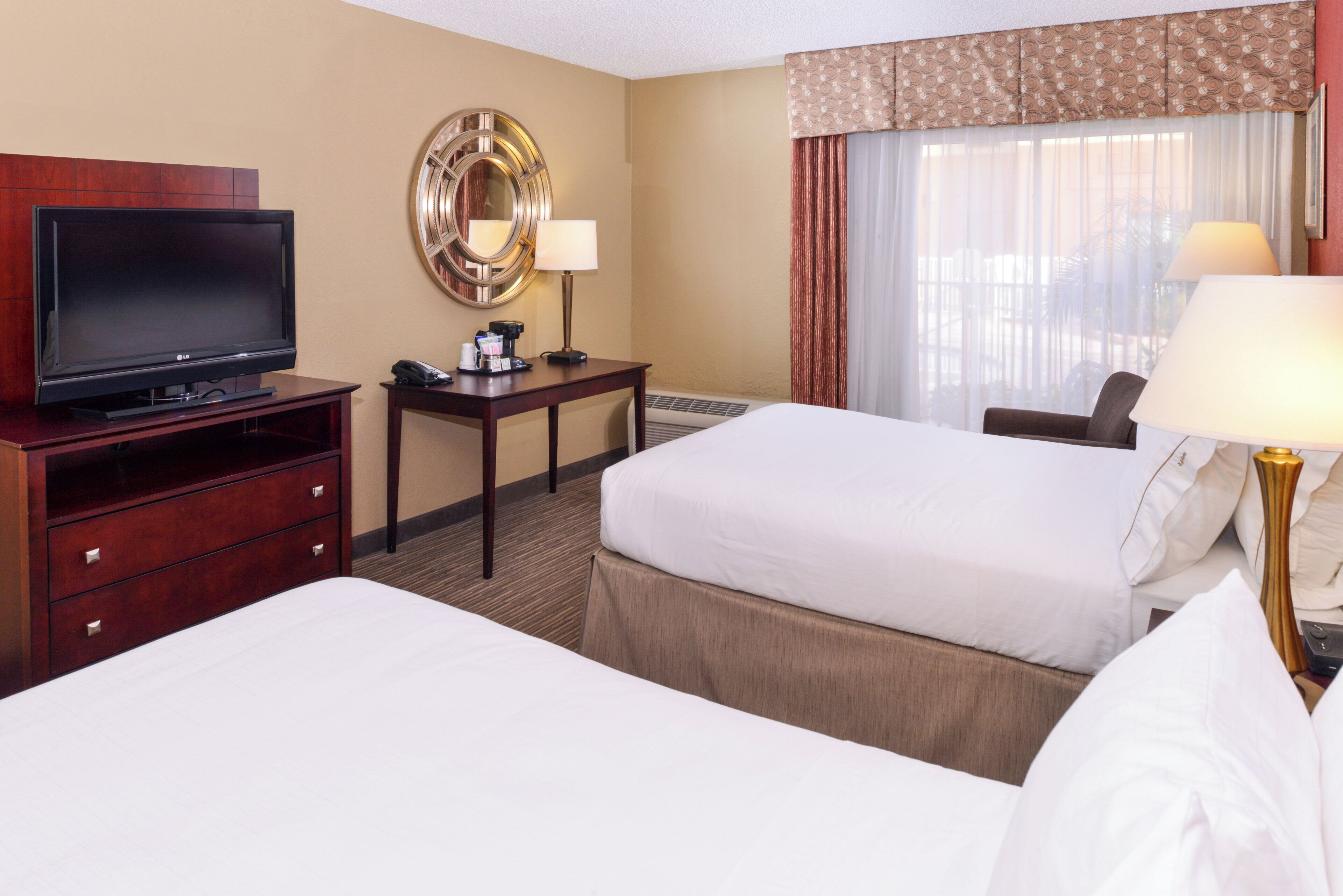 A hotel room with two beds, a TV, a desk with a lamp, and a window with curtains.