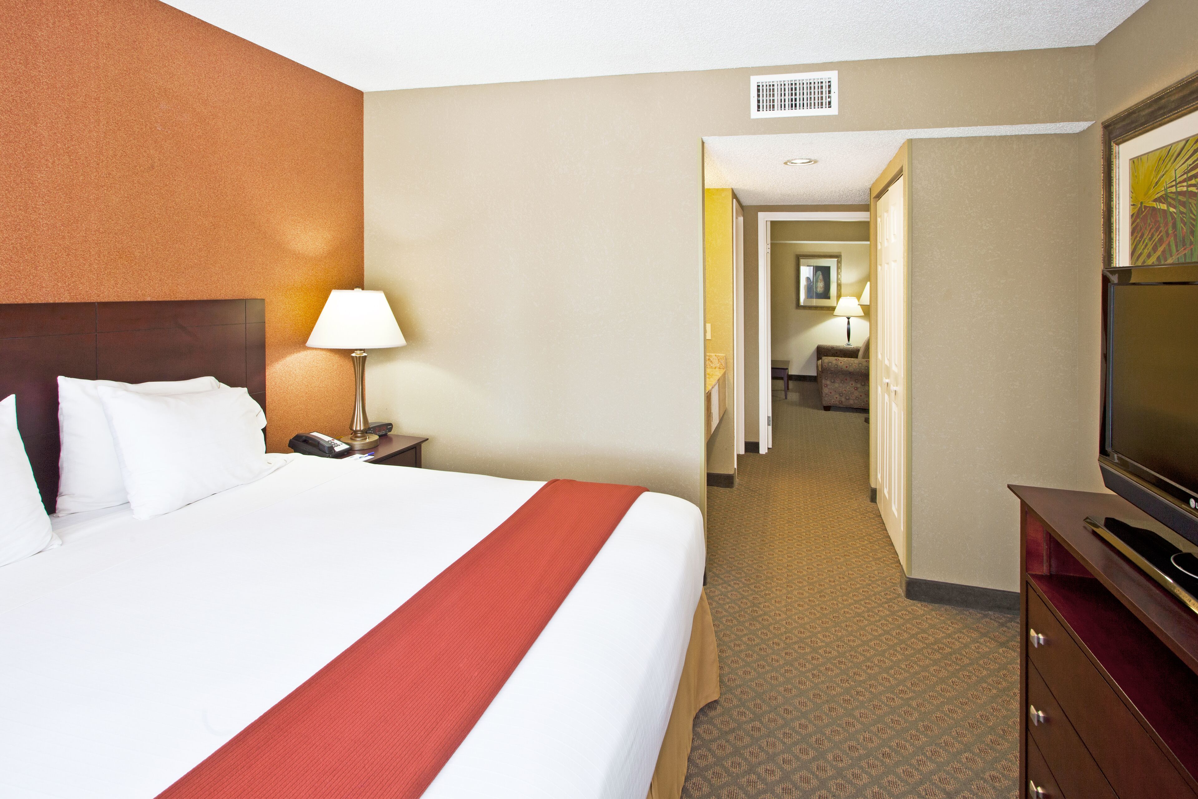 A hotel room with a large bed, a TV, a desk with a lamp, and a hallway leading to another room.