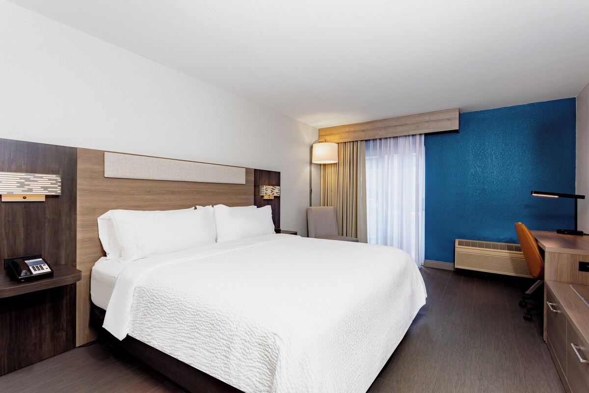 A hotel room with a large bed, a desk, and a chair. There is a nightstand with a phone and a lamp. The room has a blue accent wall and a window with curtains.