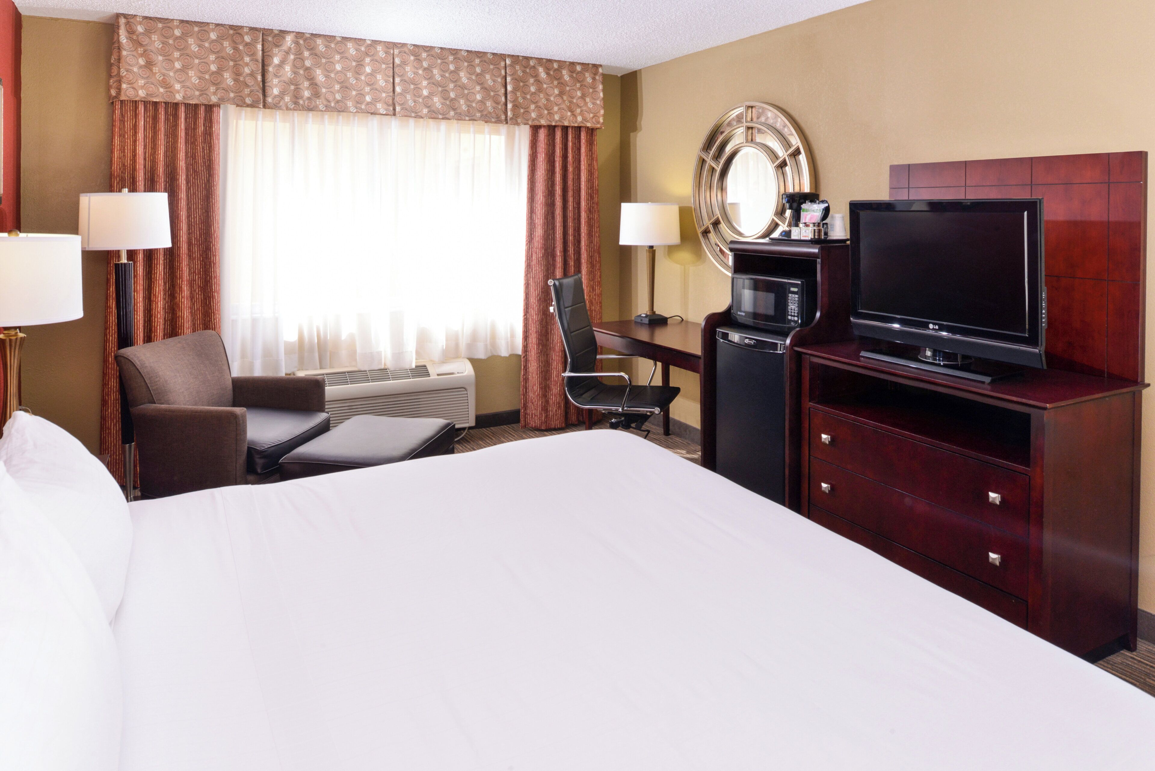 Une chambre d’hôtel avec un grand lit, un bureau équipé d’un ordinateur et d’un téléviseur à écran plat, une chaise, une lampe de bureau et une fenêtre avec des rideaux.