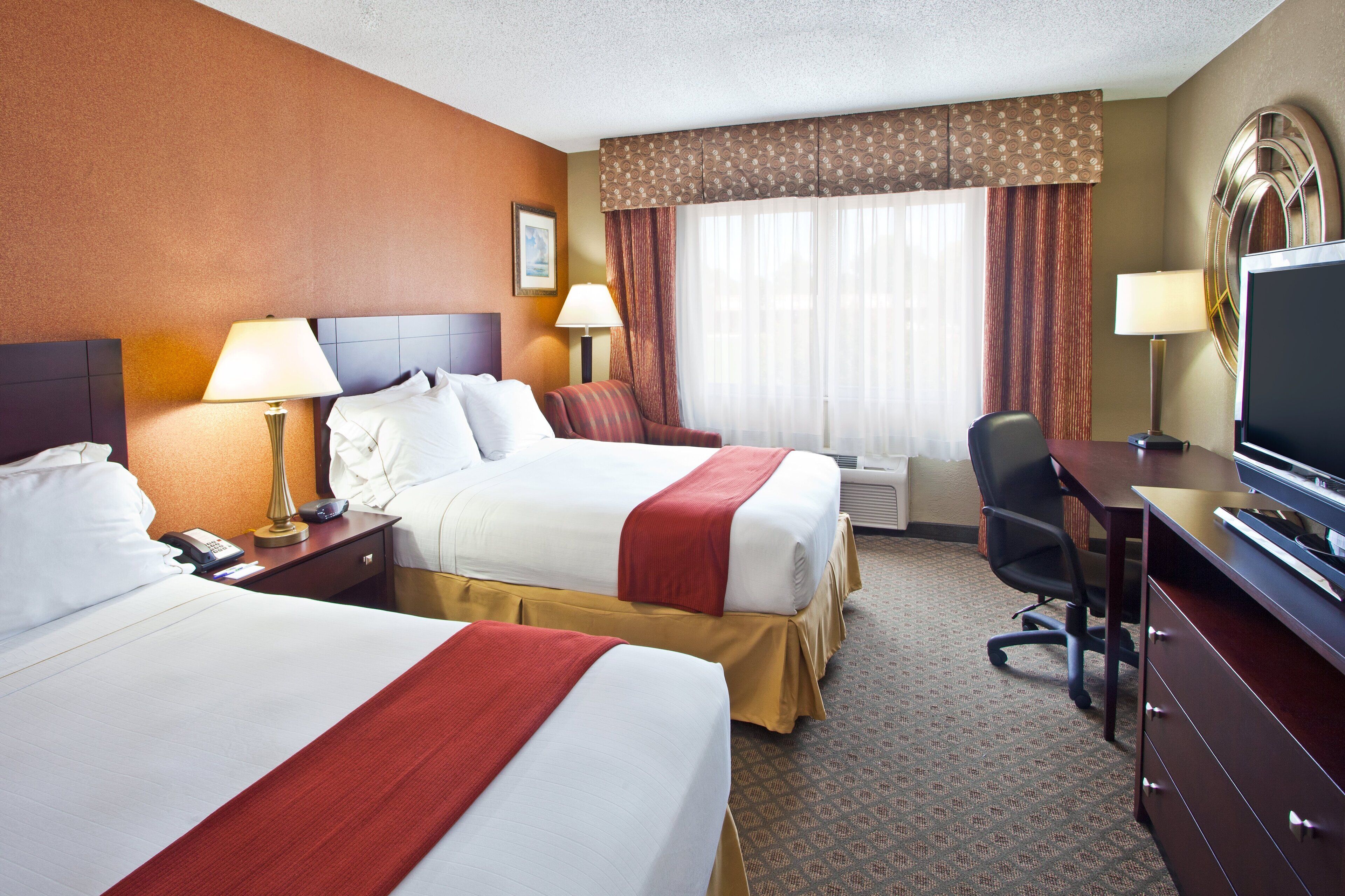 A hotel room with two beds, a desk, a TV, and a window with curtains.