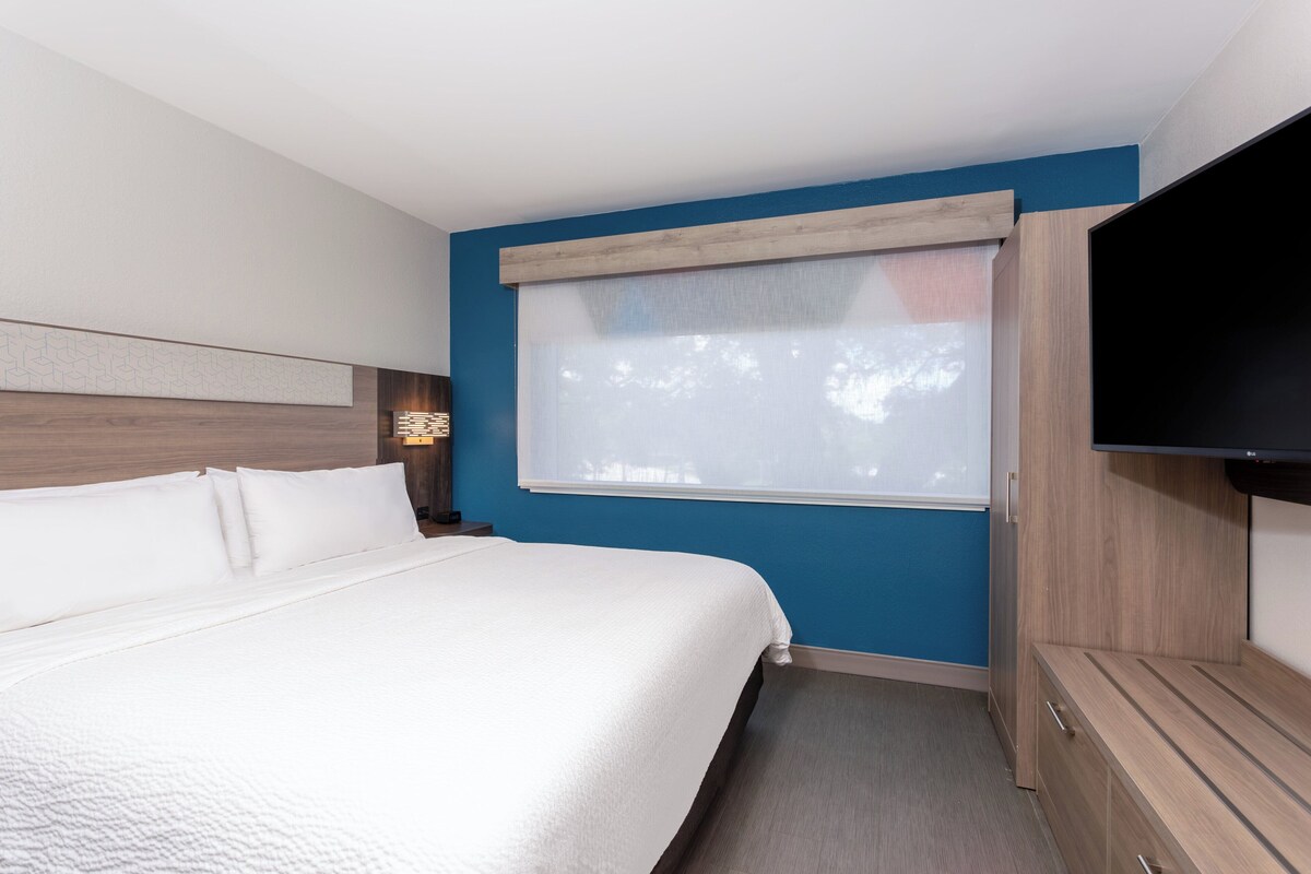 A hotel room with a large bed, a television, and a window with blinds.