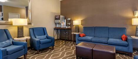 A hotel room with a blue sofa, two armchairs, a brown ottoman, a coffee machine, and a mirror on the wall.