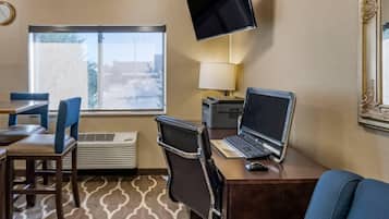 A hotel room with a desk, laptop, and TV. There is a window with a view, a chair, and a lamp.
