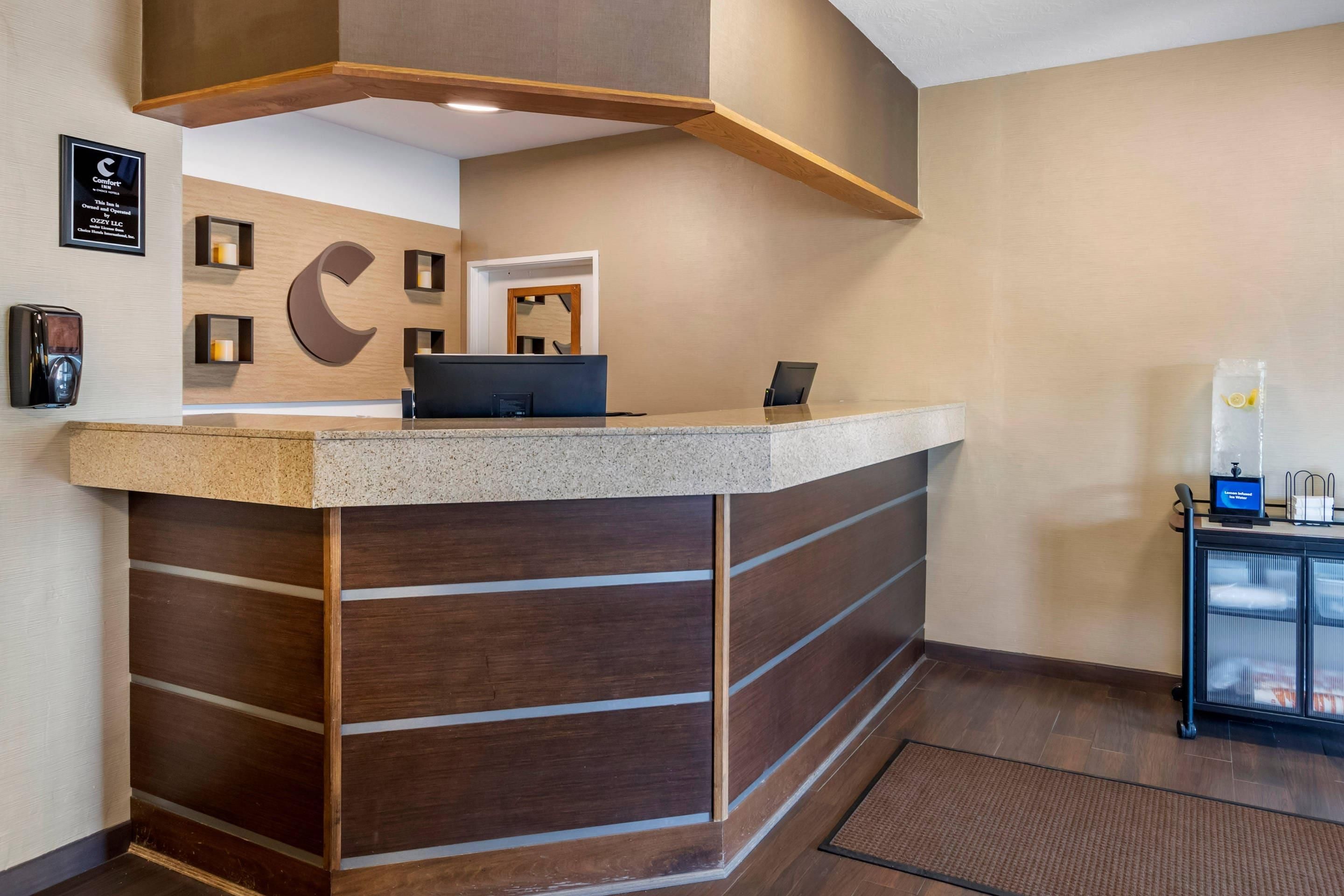 Un comptoir d’accueil avec un panneau décoratif en bois, un grand logo en forme de C sur le mur et un petit coin salon équipé d’une machine à café.