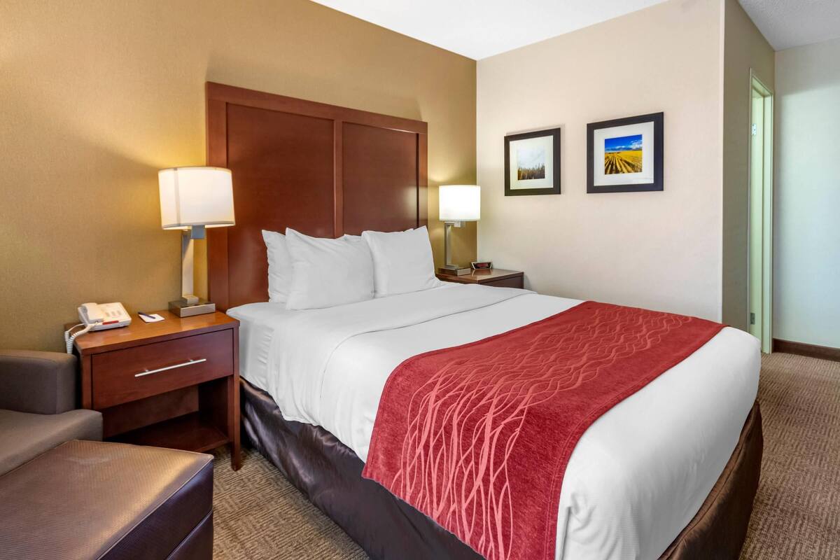 A hotel room with a large bed, a wooden headboard, a nightstand with a lamp, a desk, and a chair. There are two framed pictures on the wall.