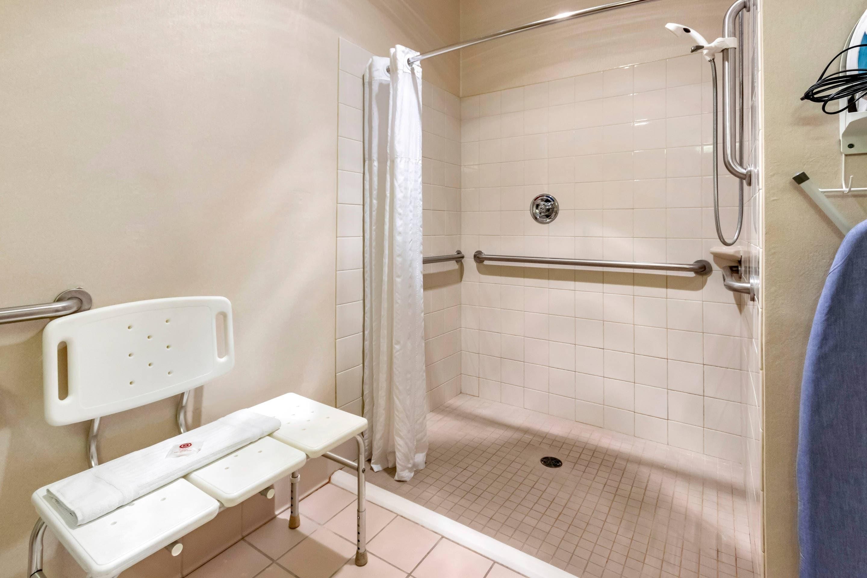 Ducha accesible en silla de ruedas con asiento, barras de apoyo y suelo de baldosas blancas.