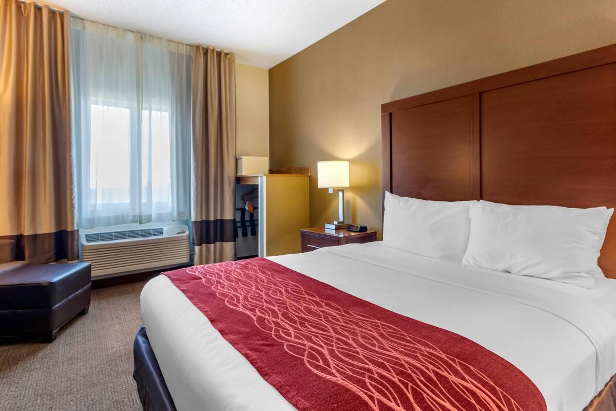 A hotel room with a large bed, a wooden headboard, a bedside table with a lamp, a small desk with a chair, and a window with curtains.