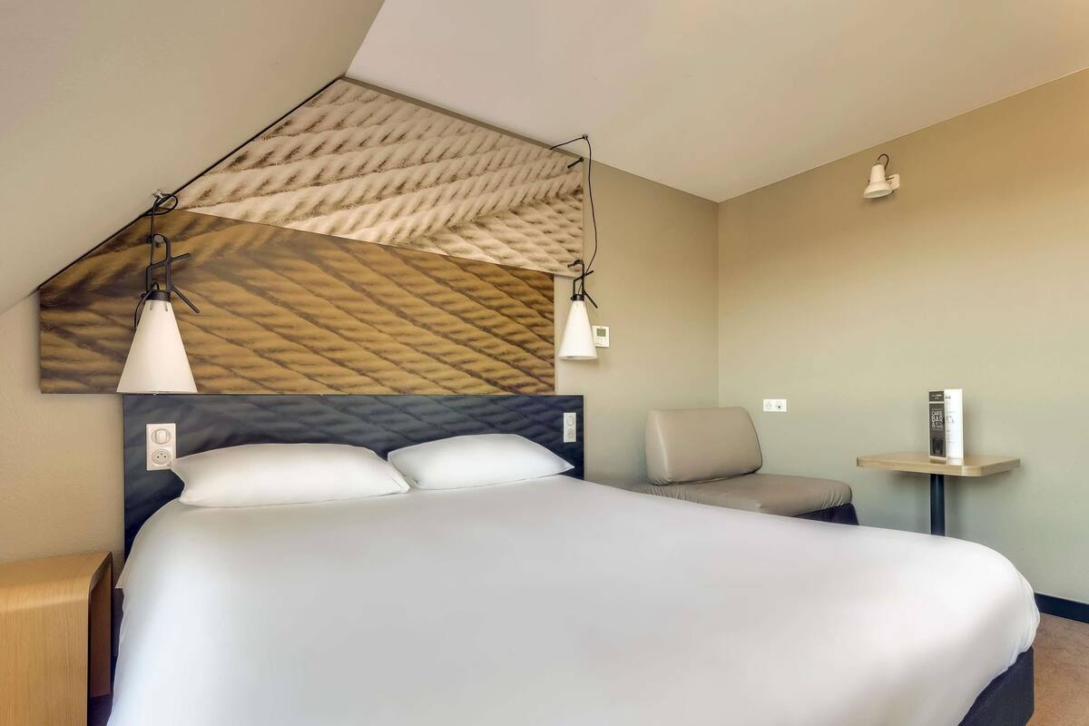 A hotel room with a large bed, two bedside tables, a chair, and a small table with a lamp.