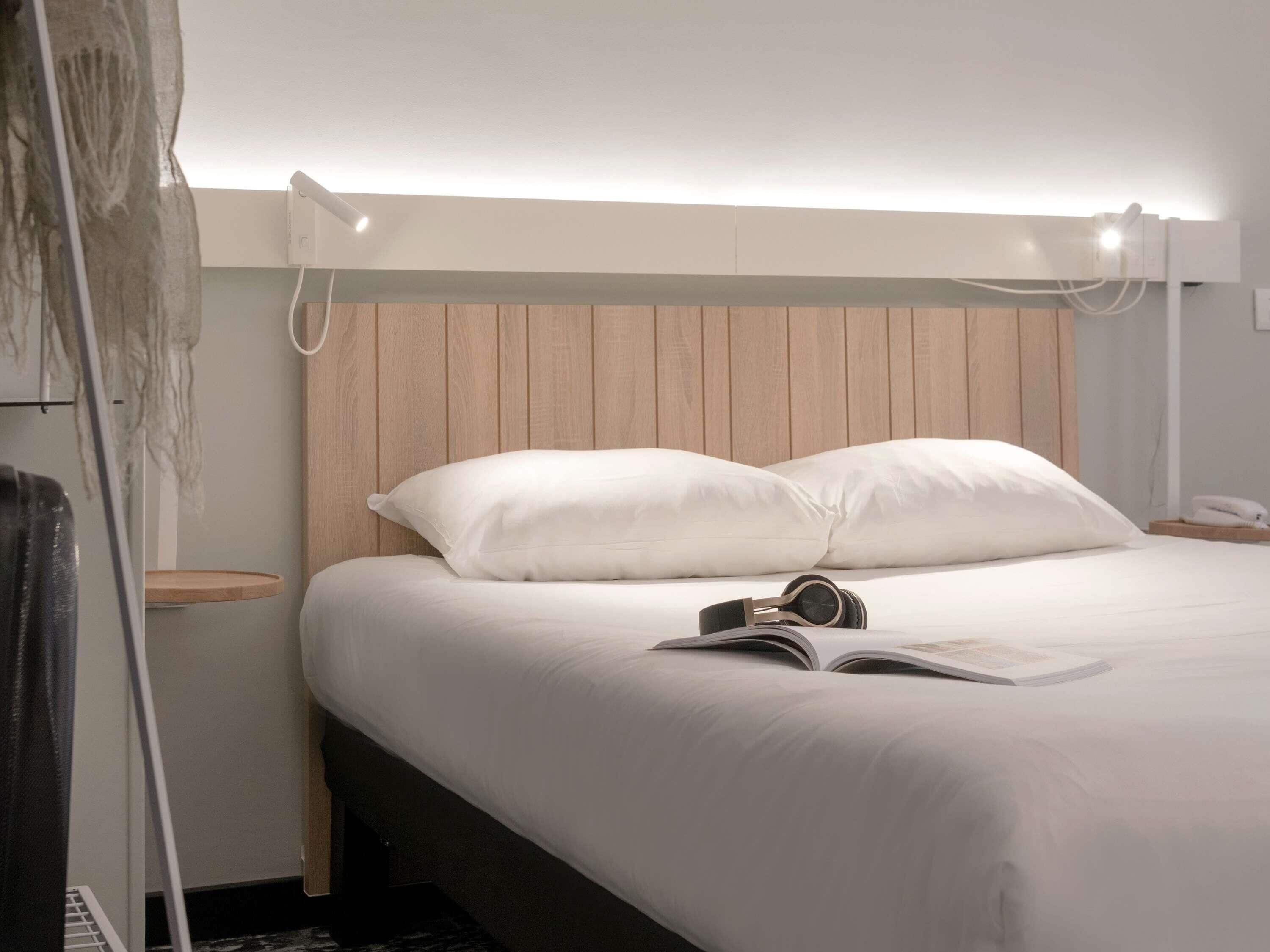 A neatly made bed with white linens, a pair of headphones, and an open book on the bedside table.