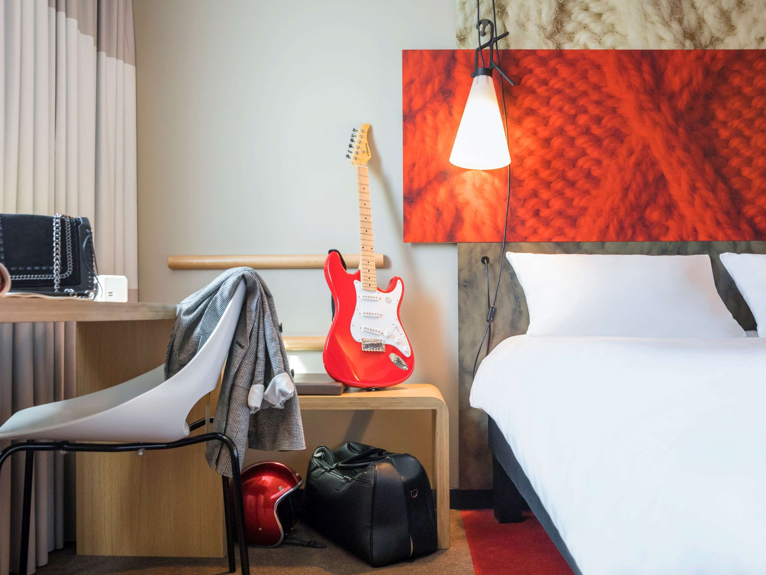 Een hotelkamer met een bed, een bureau met een stoel, een rode elektrische gitaar, een zwarte tas en een rode helm op de vloer.