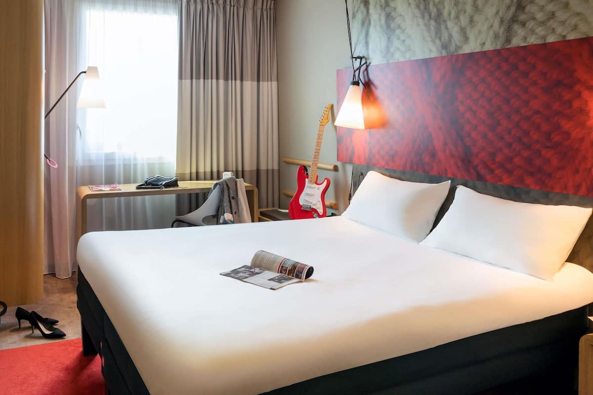 Una habitación de hotel con una cama grande, una guitarra roja, una lámpara y una revista sobre la cama.