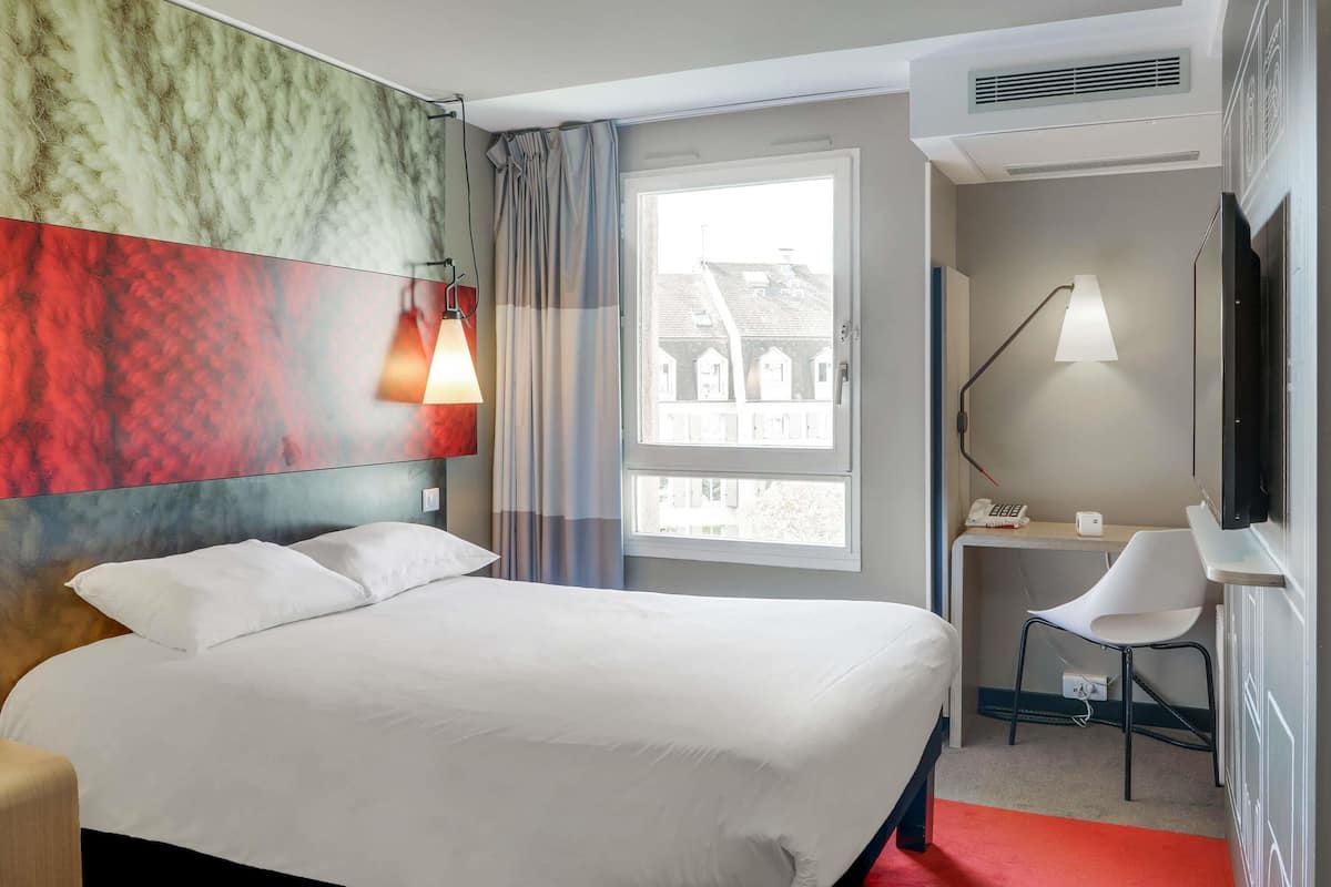 Habitación de hotel con cama, escritorio, silla, televisor y ventana con vista a edificios.