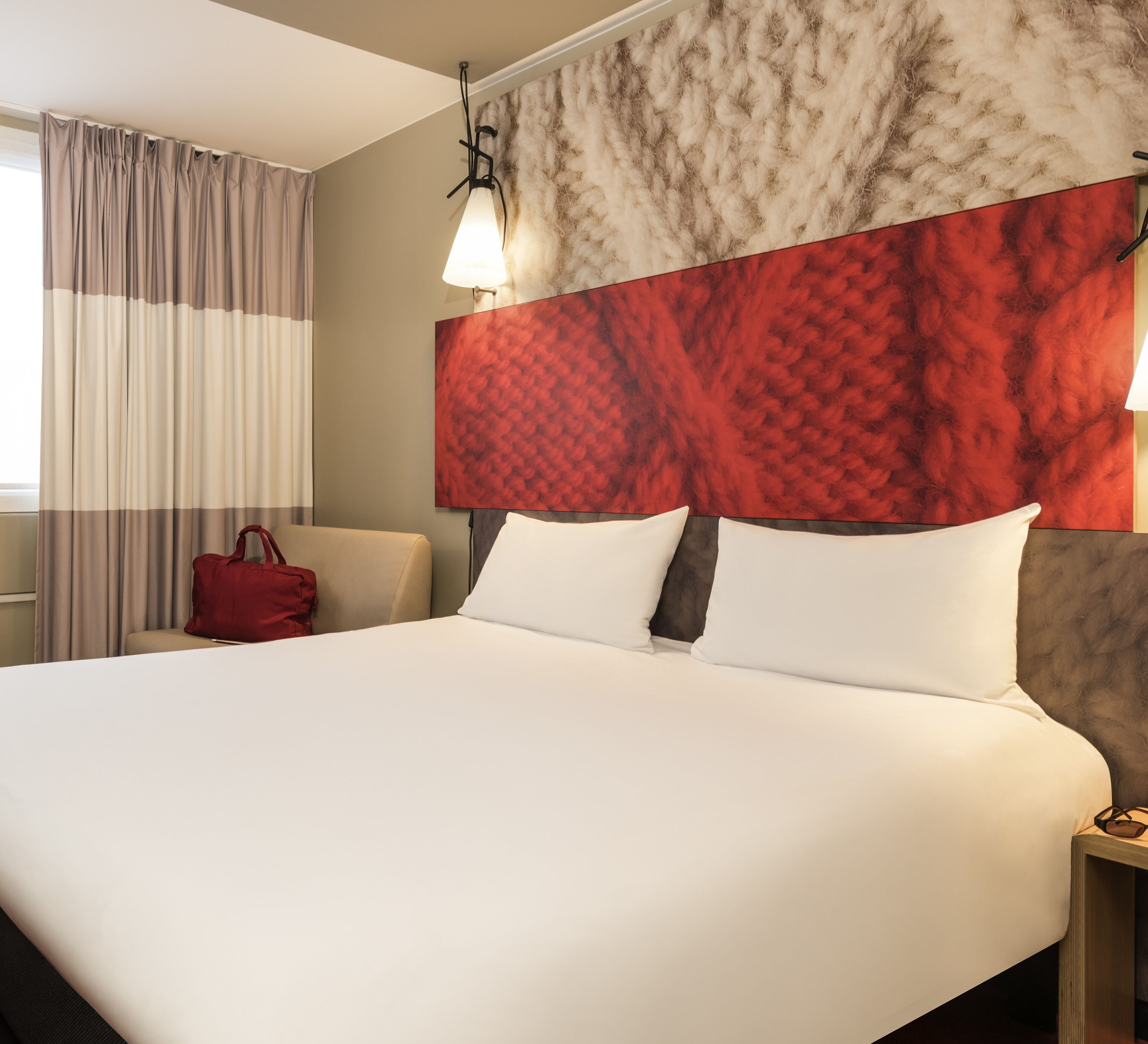 A hotel room with a large bed, a red handbag on the armchair, and a red and beige textured wall.