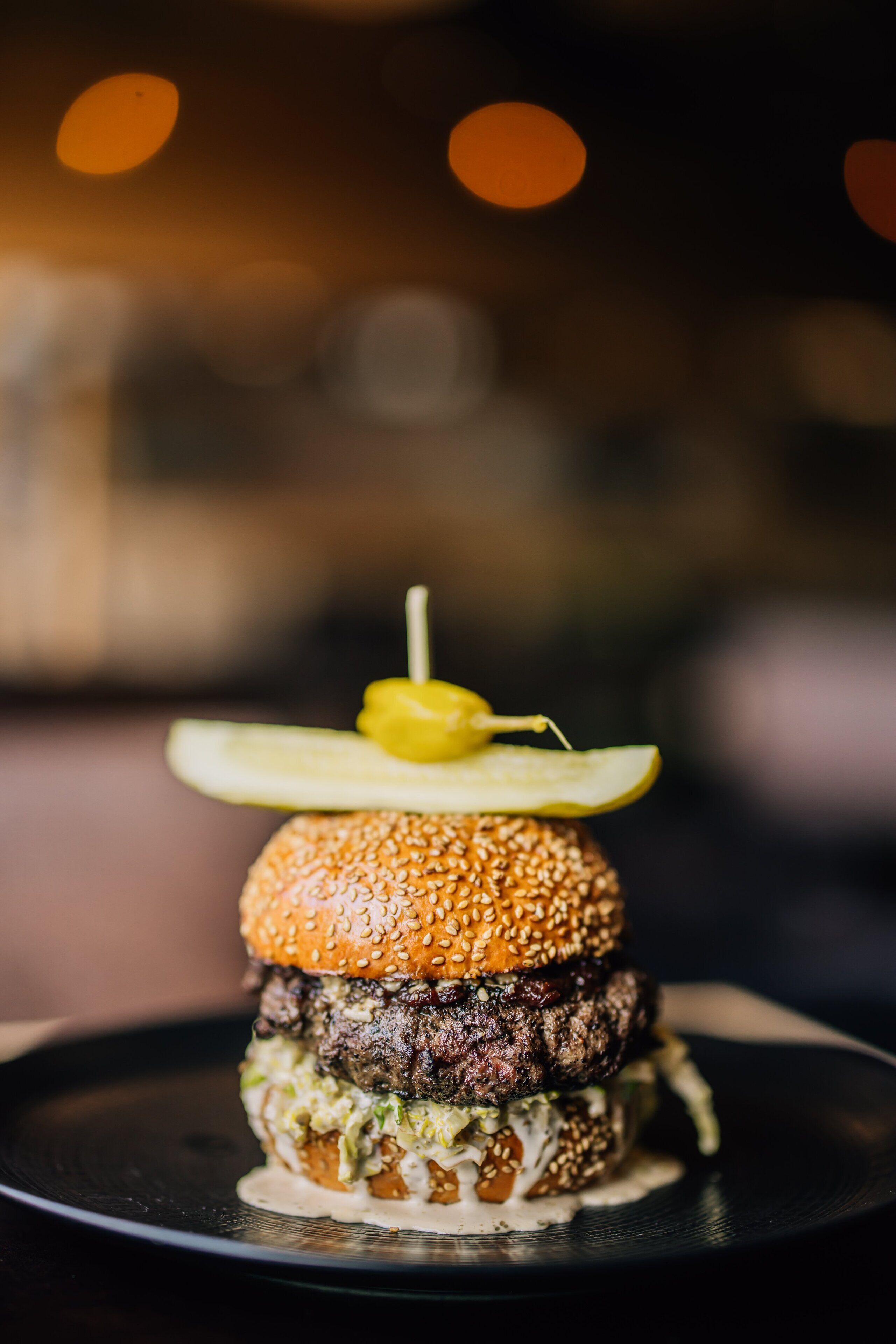 Close-up van een gourmet hamburger met een augurk erop, geserveerd op een zwart bord.