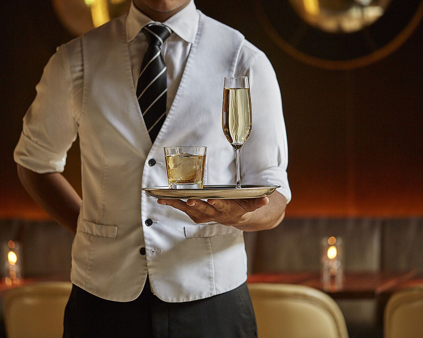 A person in a white shirt and tie holding a tray with a glass of champagne and a small glass of liquor.