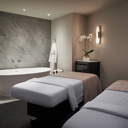 A modern bathroom with a freestanding bathtub, two beds with white bedding, a small table with a candle, and a wall-mounted TV.