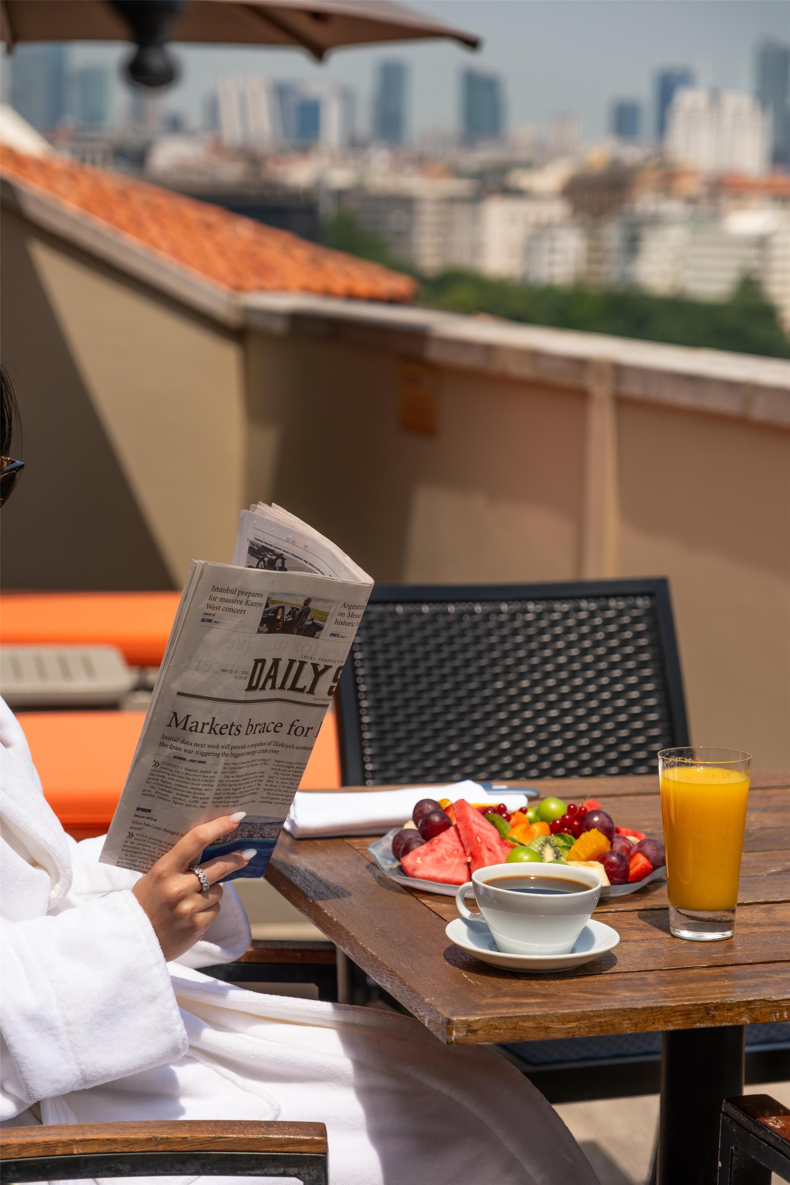 A person reading a newspaper at a table with a city view, fruit, and a glass of orange juice.