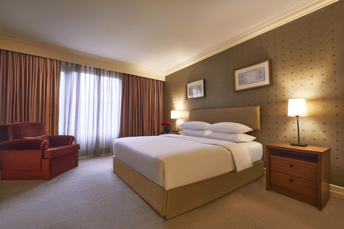 A hotel room with a bed, a chair, a nightstand, a lamp, and a window with curtains.