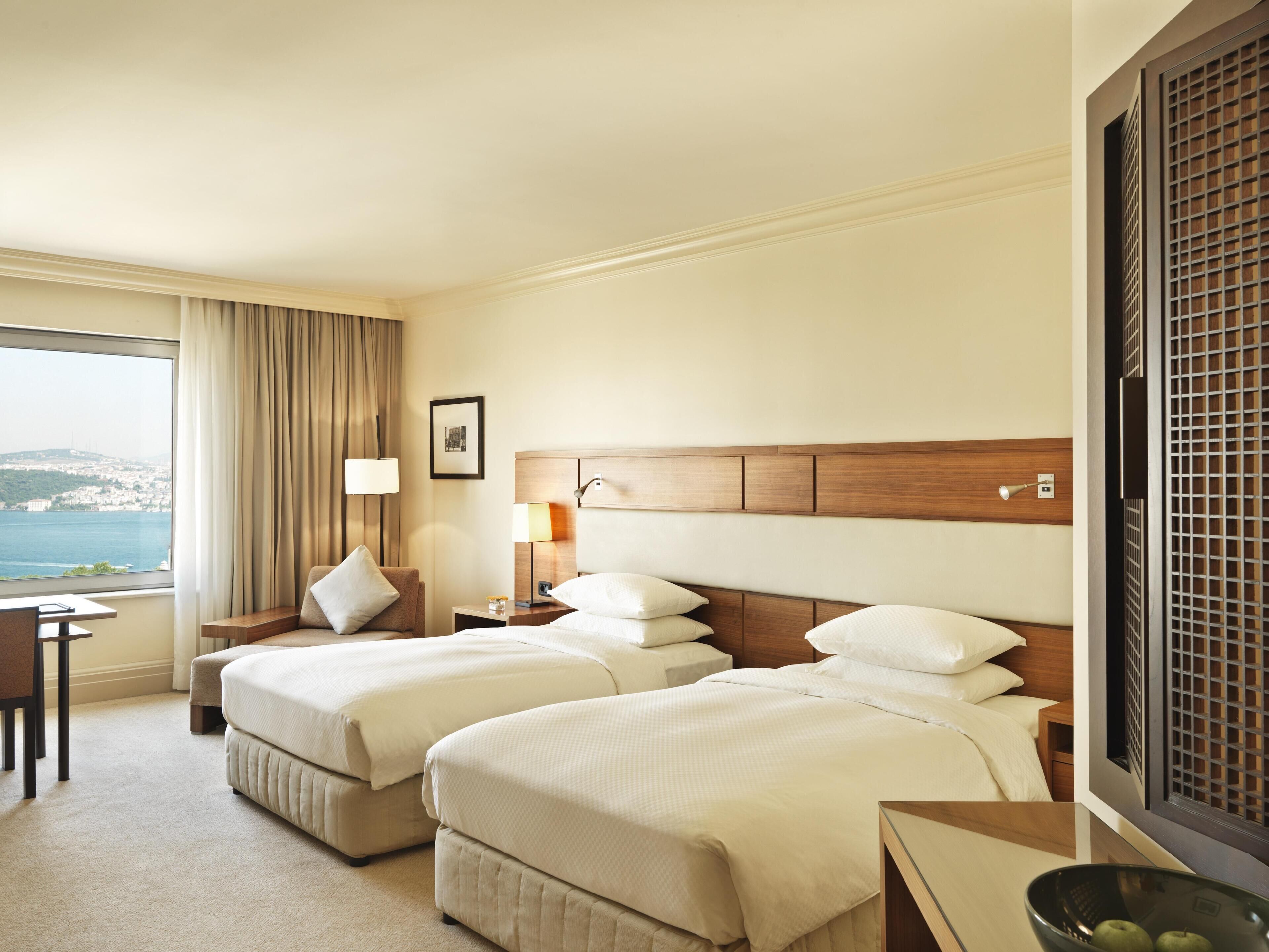 A hotel room with two beds, a desk, a chair, a lamp, and a view of the sea.