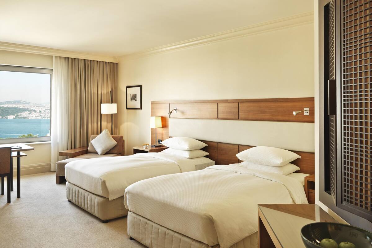 A hotel room with two beds, a desk, a chair, a lamp, and a view of the sea.
