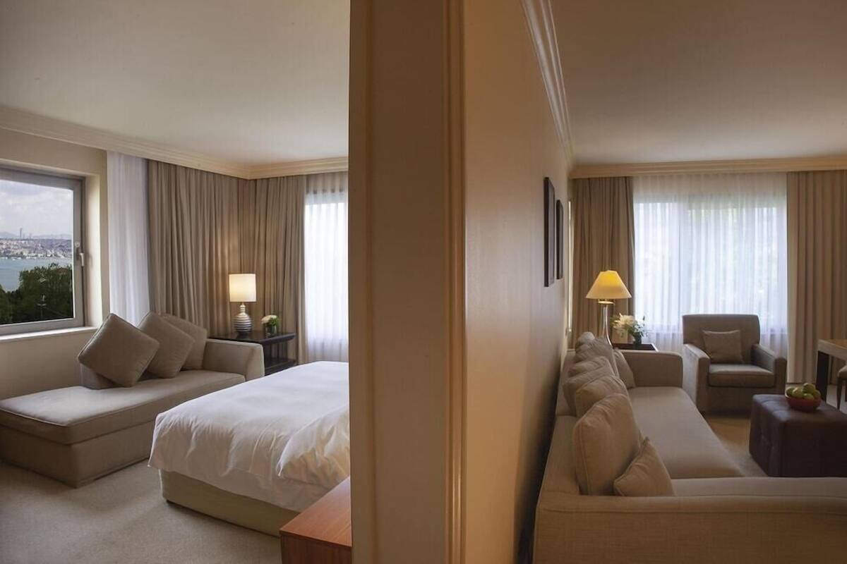 A hotel room with a bed, two sofas, a chair, a lamp, and a view of the city.