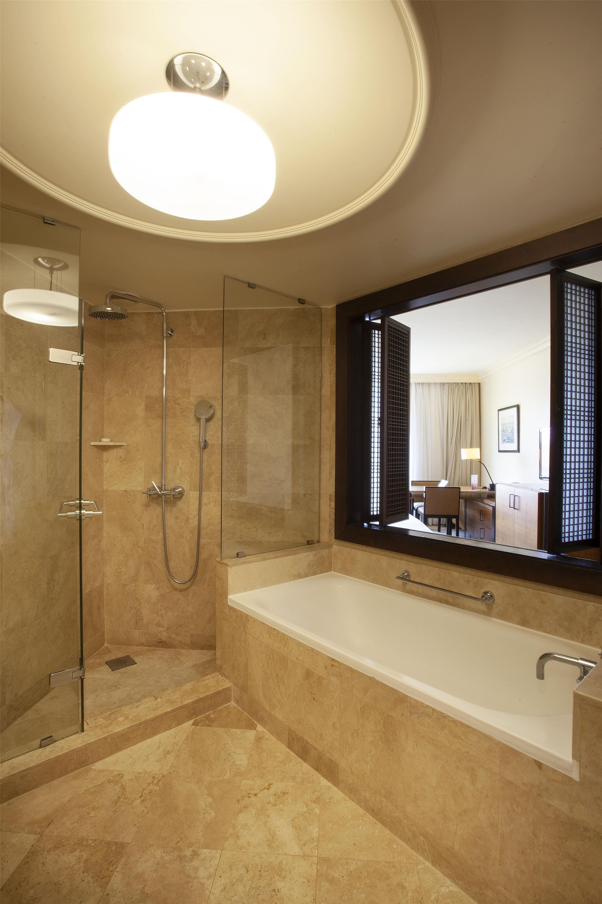 Un baño moderno con ducha envuelta en vidrio, una bañera grande y un espejo que refleja un cuarto con escritorio y silla.