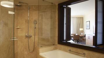 Un baño moderno con ducha envuelta en vidrio, una bañera grande y un espejo que refleja un cuarto con escritorio y silla.