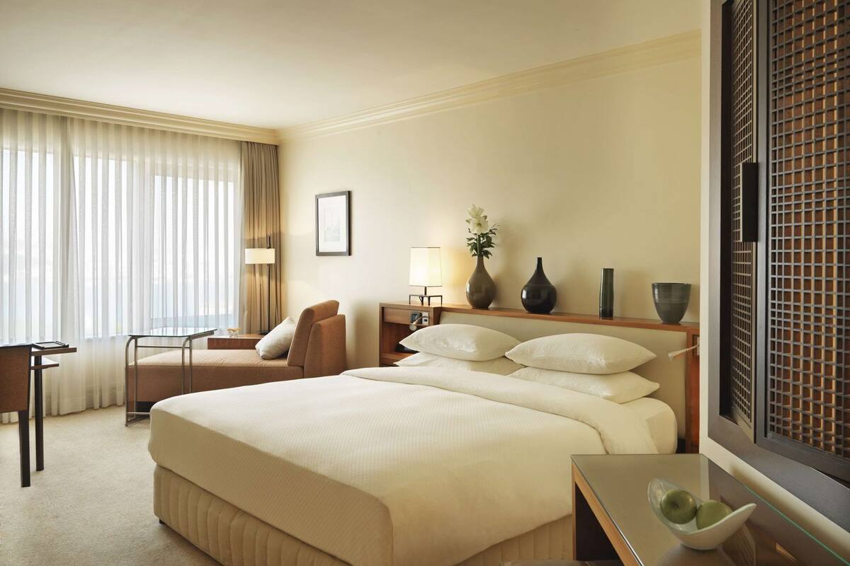 A hotel room with a large bed, a desk, a chair, a nightstand with lamps, and a window with curtains.