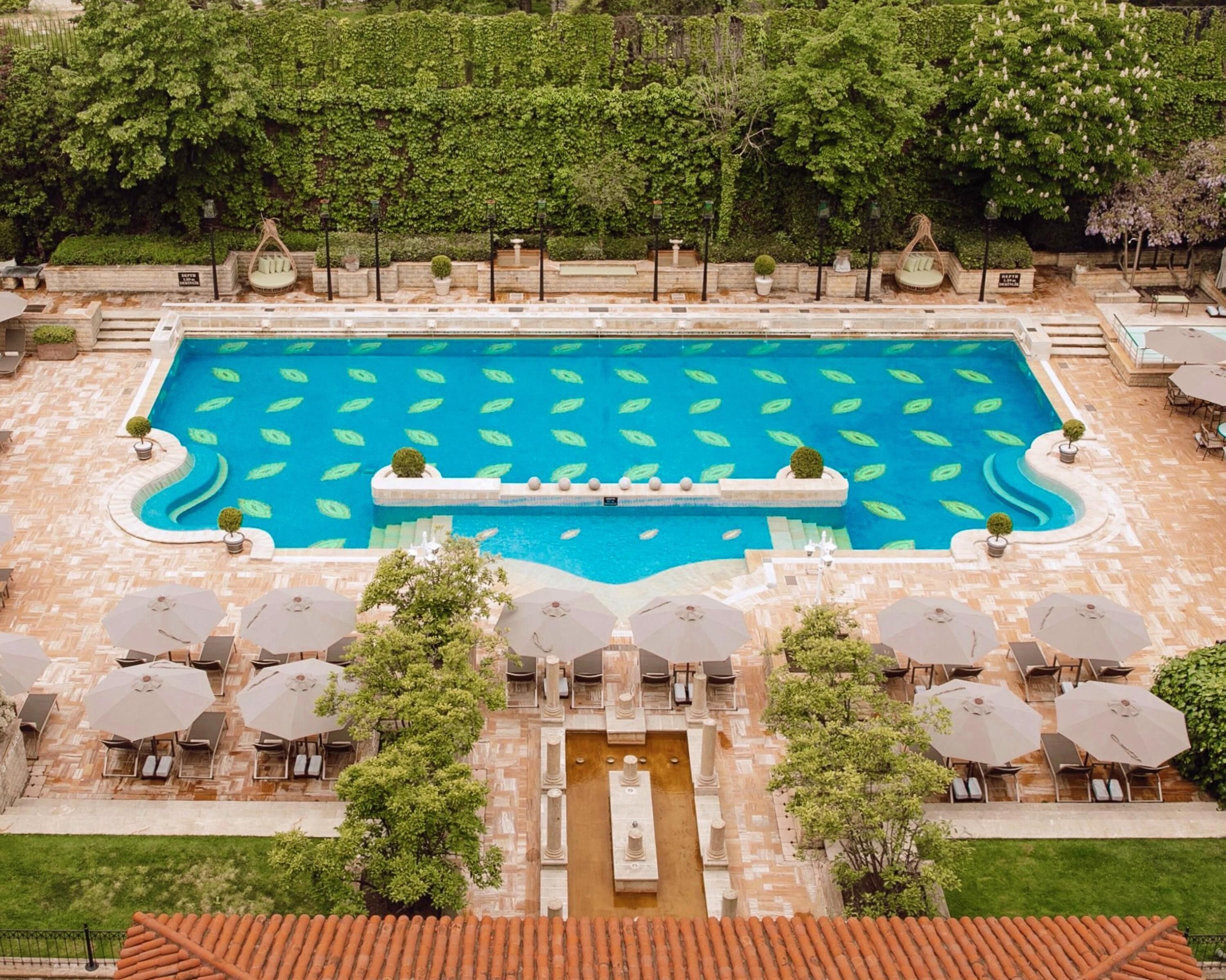 Ein großer, rechteckiger Pool mit Blattmuster, umgeben von Liegestühlen und Sonnenschirmen, mit einem Steinpfad, der zu einem kleinen Brunnen führt.