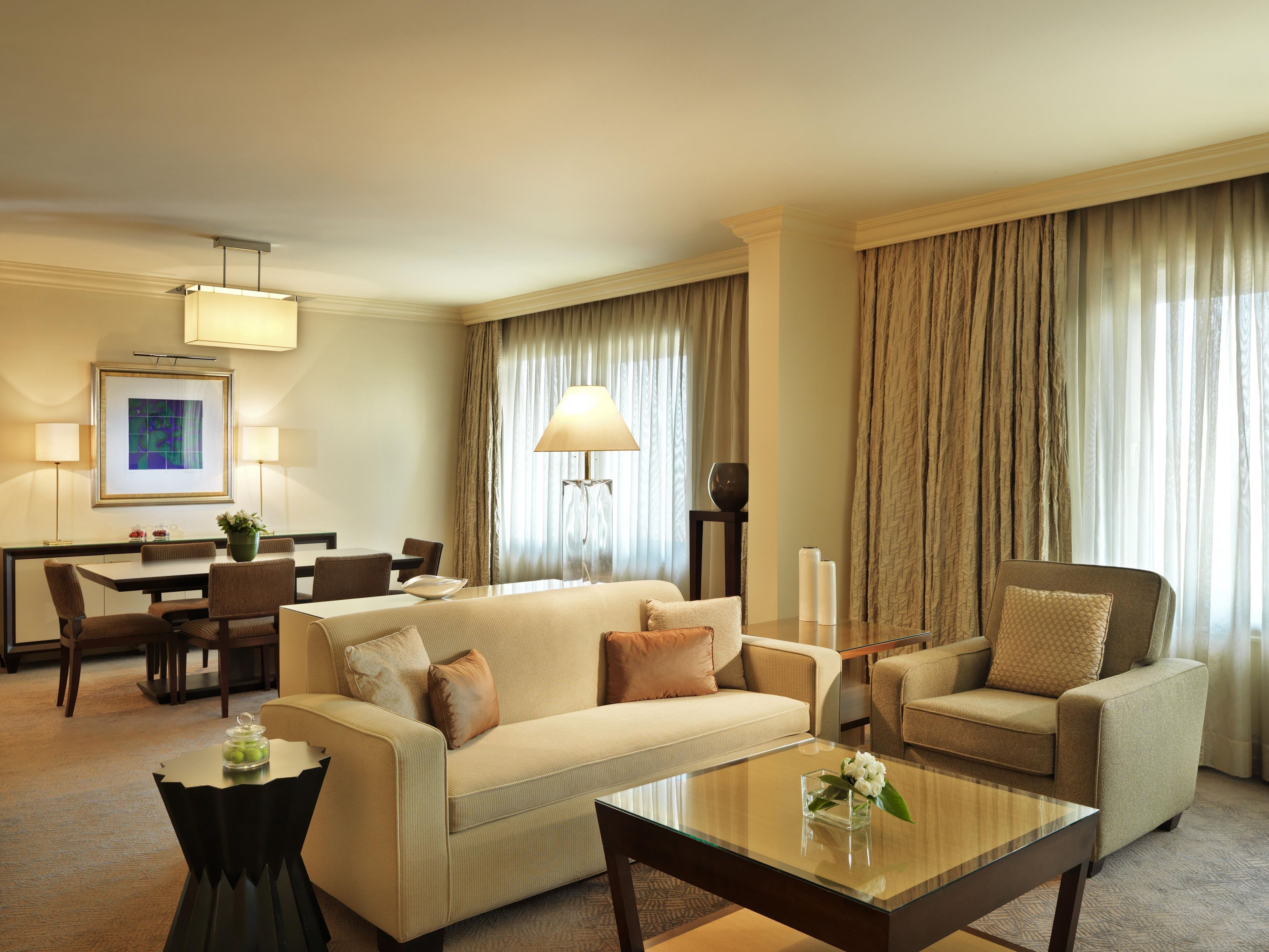 A hotel room with a sofa, armchair, dining table, and chairs. There is a lamp, a vase with flowers, and a framed picture on the wall.