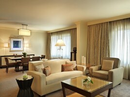A hotel room with a sofa, armchair, dining table, and chairs. There is a lamp, a vase with flowers, and a framed picture on the wall.