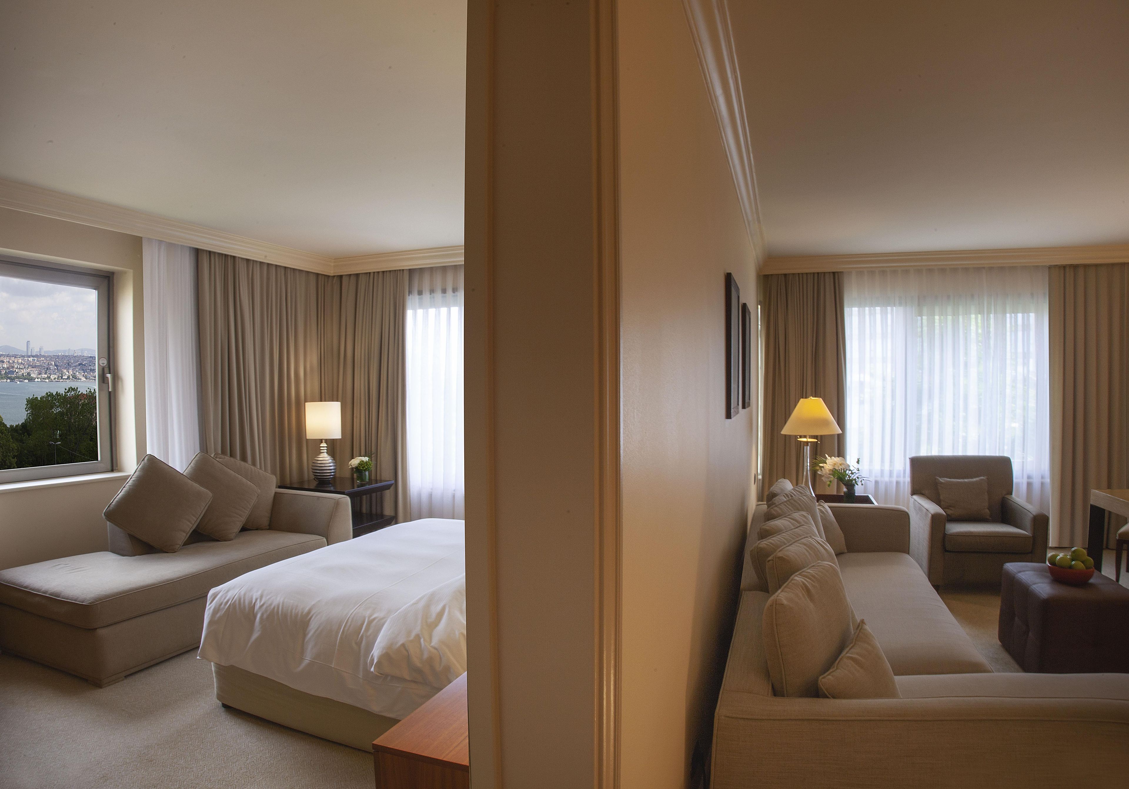 Habitación de hotel con cama, sofá, sillón, lámpara y vista a la ciudad.