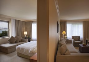 Habitación de hotel con cama, sofá, sillón, lámpara y vista a la ciudad.