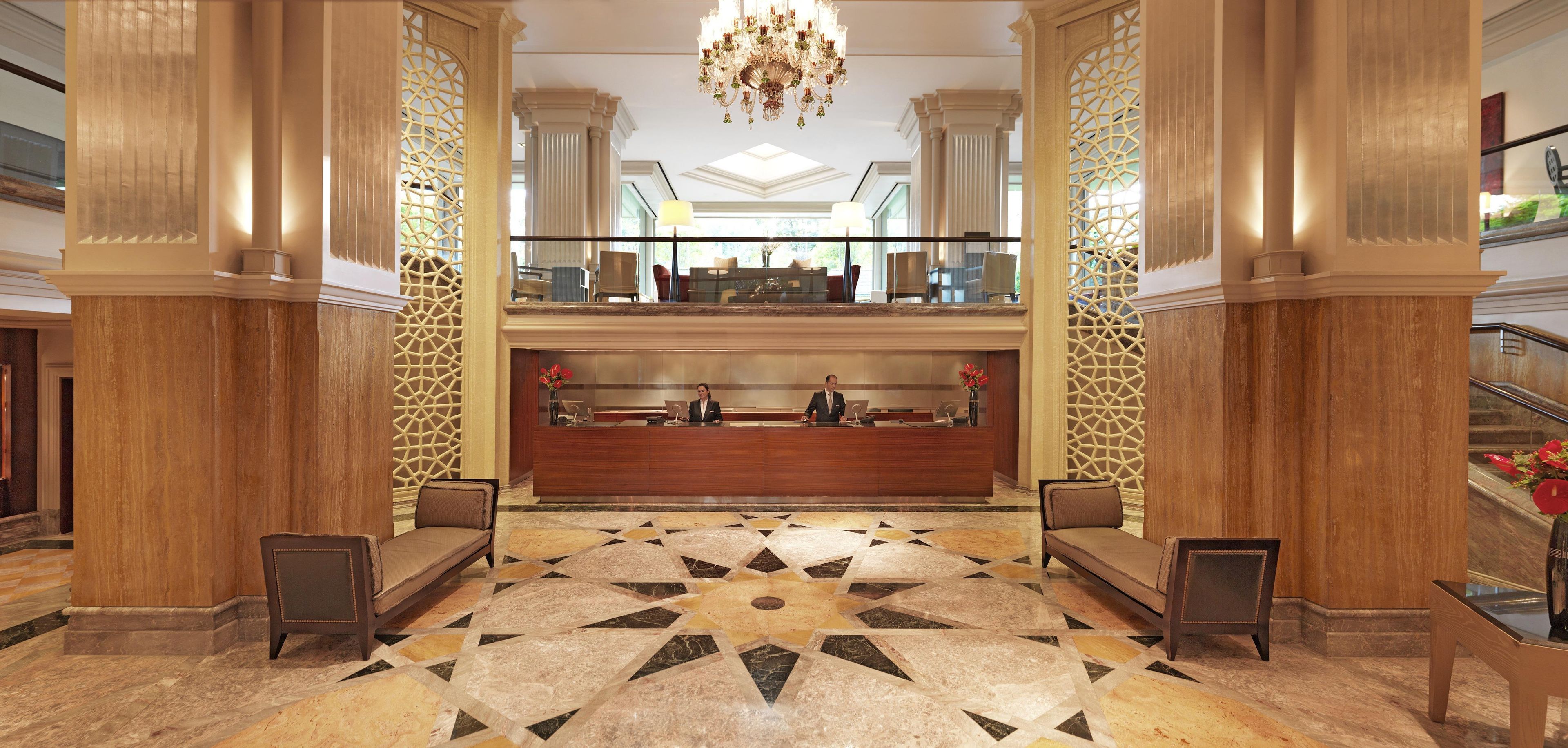 A hotel lobby with a reception desk, two sofas, a chandelier, and a patterned floor.
