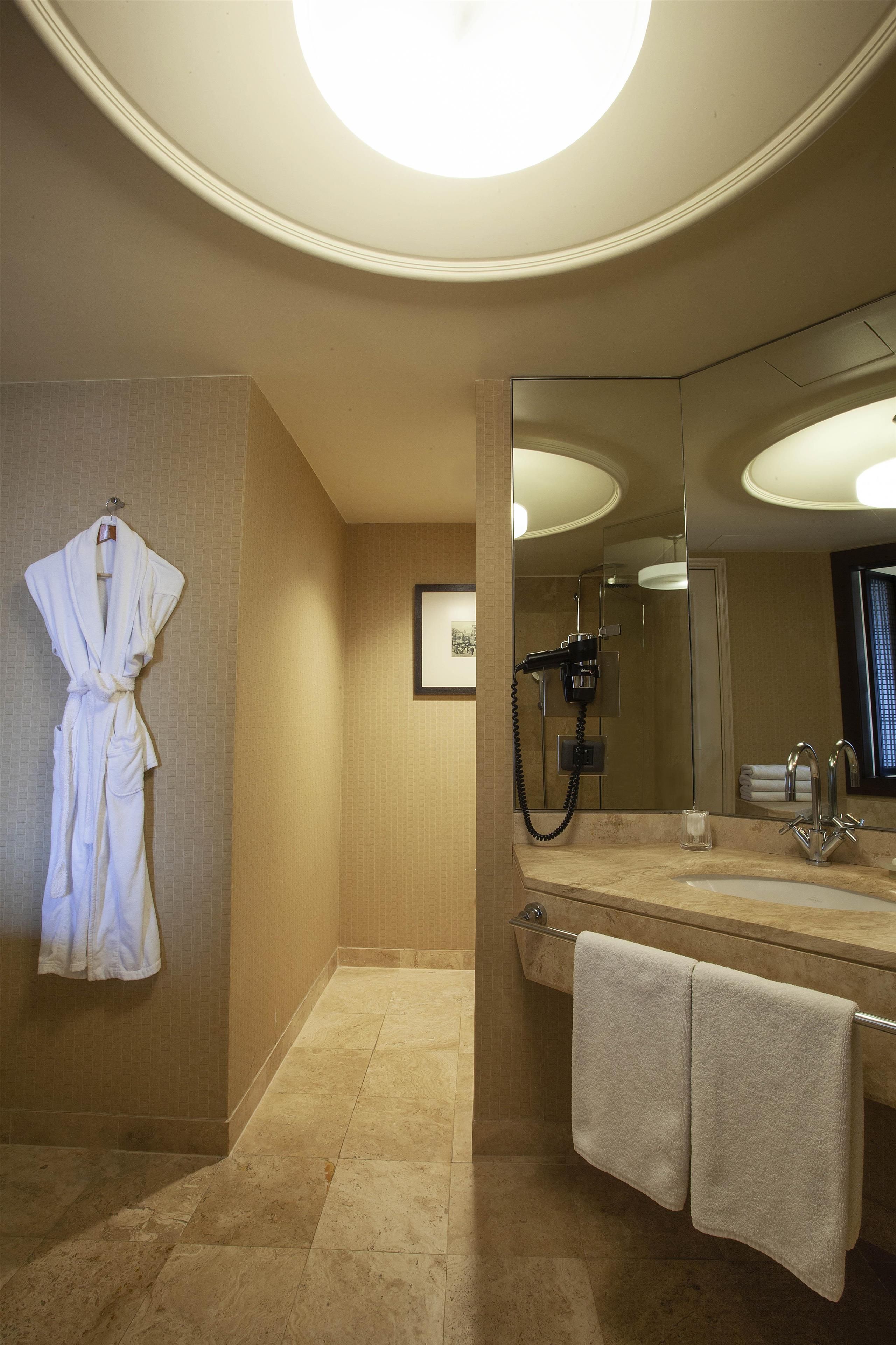 Un bagno con un accappatoio appeso al muro, un portascopevoli con asciugamani e una zona doccia, oltre a uno specchio grande.