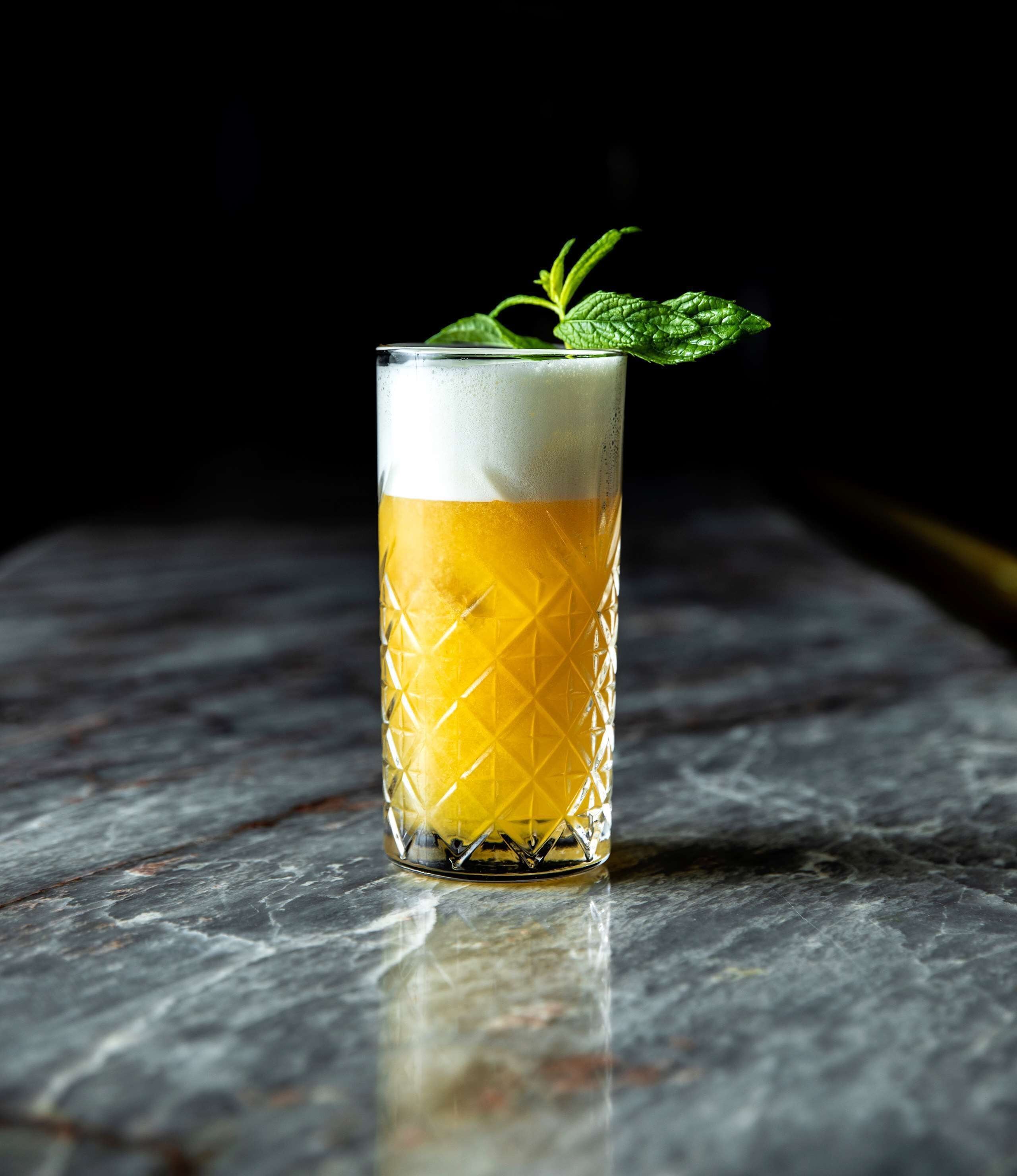 A tall glass with a textured pattern, filled with a yellow liquid and topped with a white foam, placed on a marble surface.