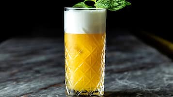 A tall glass with a textured pattern, filled with a yellow liquid and topped with a white foam, placed on a marble surface.