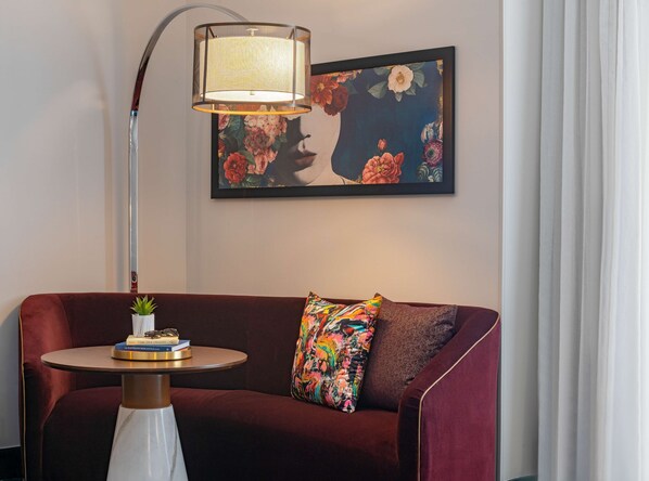 Una habitación con un sofá rojo, una mesa redonda con libros y una planta, una lámpara de pie y un cuadro floral en la pared.